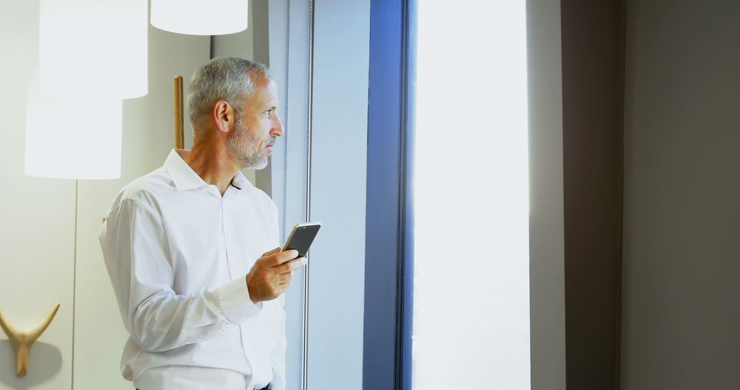 Senior Businessman Holding Smartphone Looking Out of Window - Free Images, Stock Photos and Pictures on Pikwizard.com