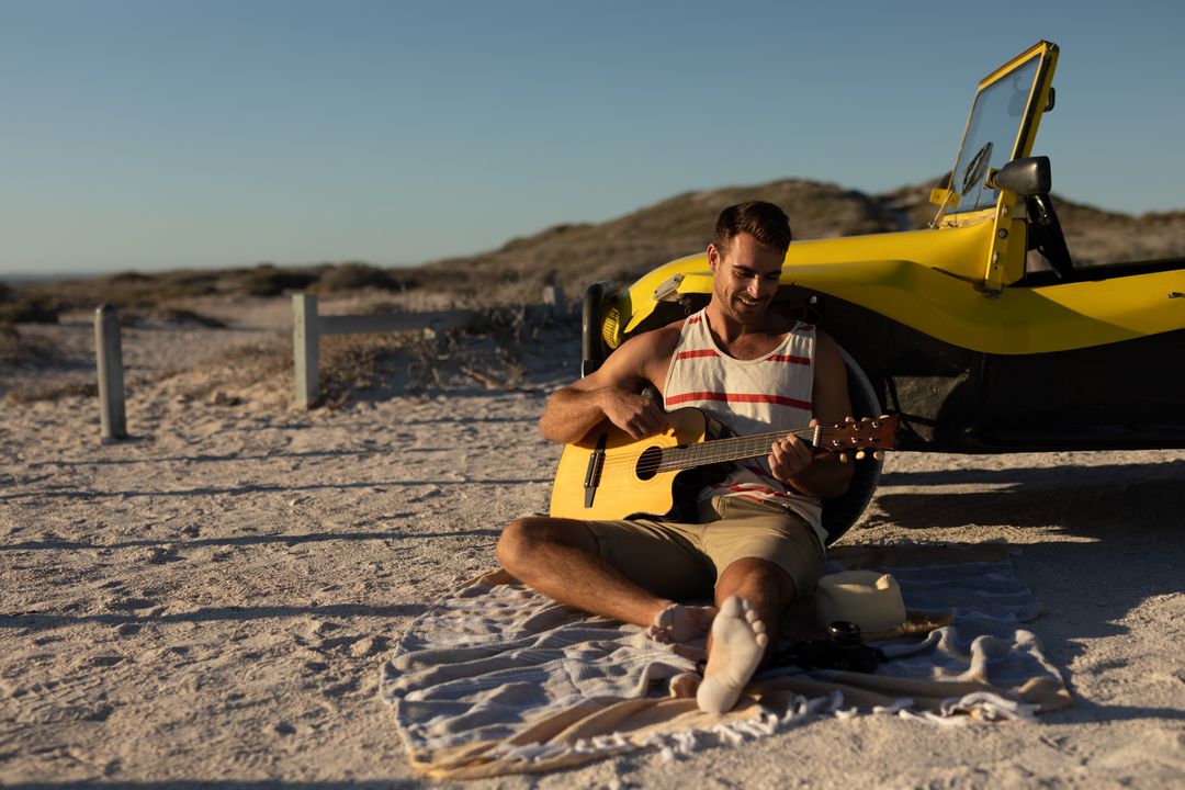 Caucasian man sitting on beach by beach buggy at sundown playing guitar - Free Images, Stock Photos and Pictures on Pikwizard.com