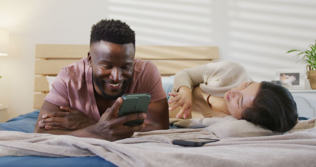 Happy diverse couple using smartphone and lying in bedroom - Free Images, Stock Photos and Pictures on Pikwizard.com