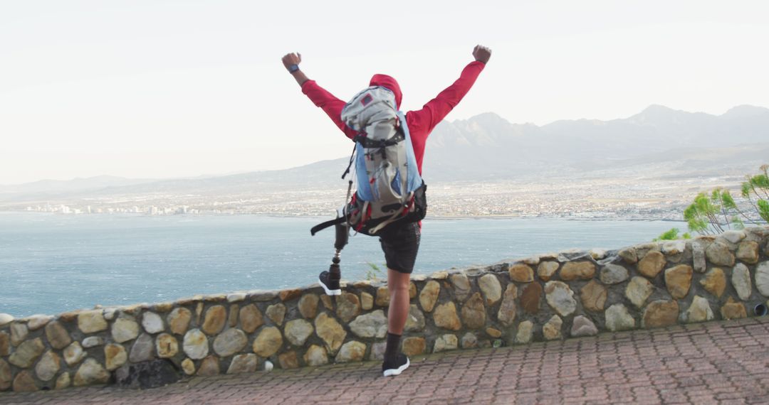Rear view of biracial man with prosthetic leg trekking, raising arms in victory looking out to sea - Free Images, Stock Photos and Pictures on Pikwizard.com