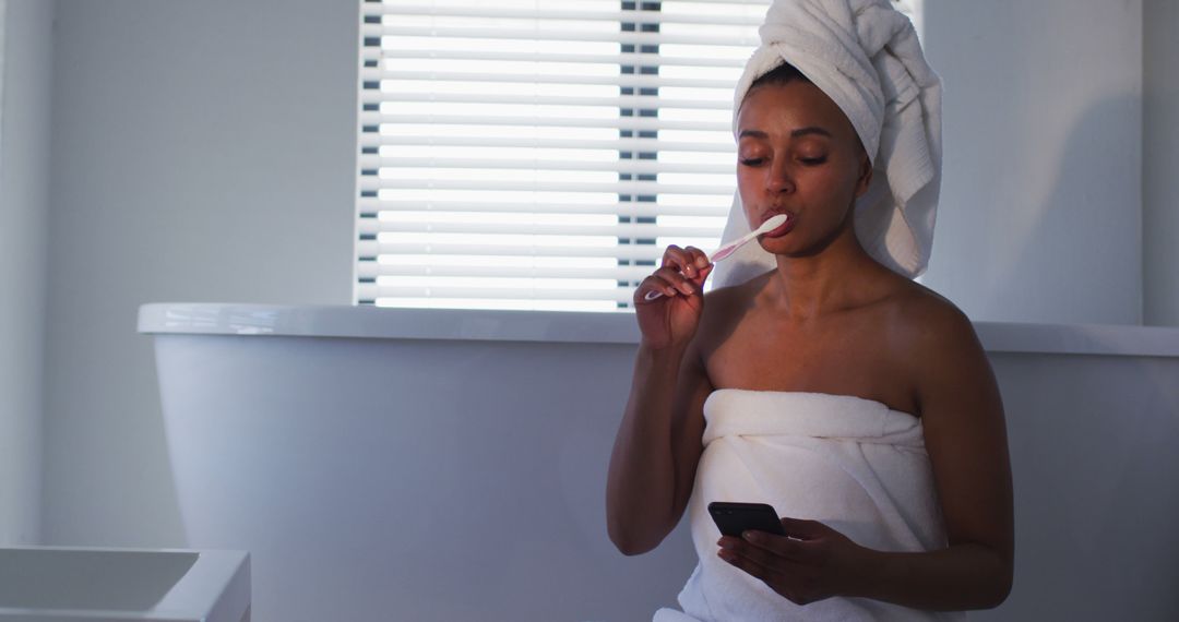 Woman Brushing Teeth and Using Smartphone in Bathroom with Towel Wrapped Around - Free Images, Stock Photos and Pictures on Pikwizard.com