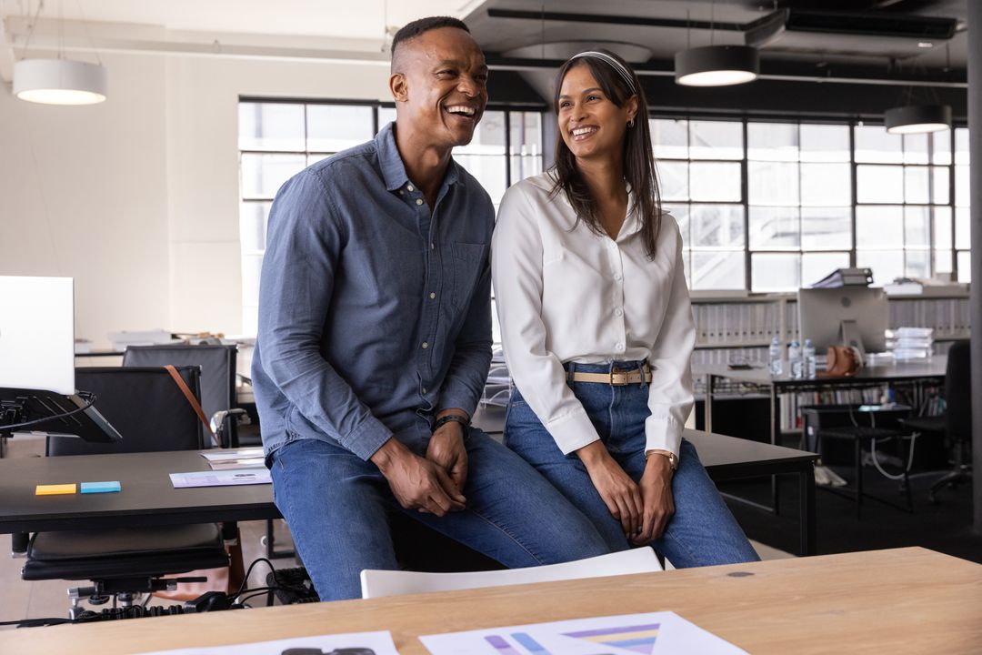 Smiling diverse coworkers sitting on desk in modern open-plan office workspace collaborating