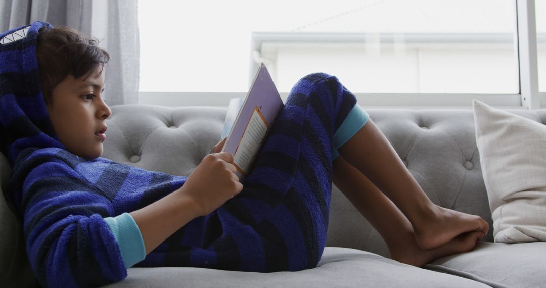 Child in Blue Striped Hoodie Relaxing on Couch Reading a Book - Free Images, Stock Photos and Pictures on Pikwizard.com