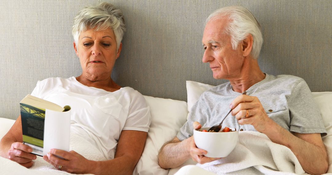 Senior Couple Relaxing in Bed Reading and Eating Breakfast