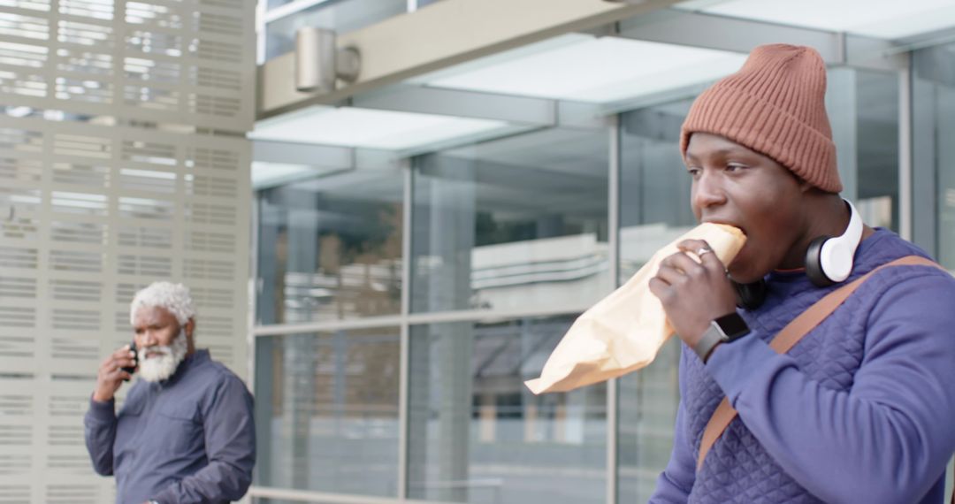 Young African American Man Eating Wrap While Older Man Talking on Phone Near Transit Hub