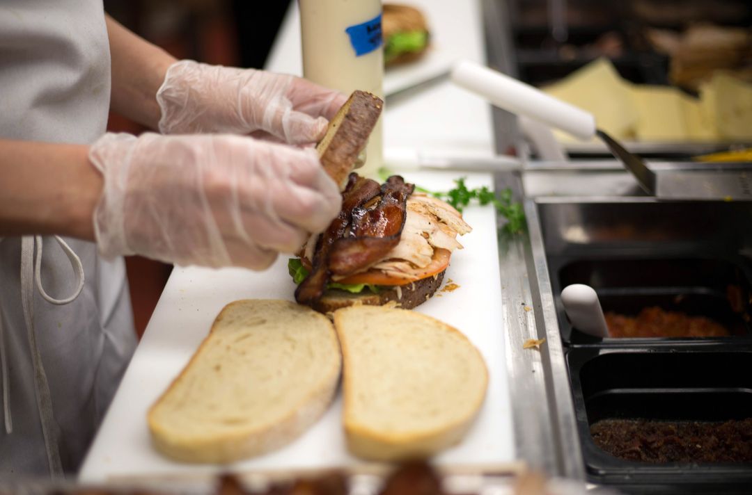 Preparing gourmet turkey bacon sandwich at deli counter with gloved hands and fresh bread