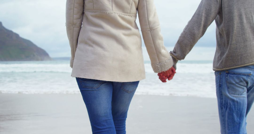 A young Caucasian couple holds hands while walking along a scenic beach, with copy space - Free Images, Stock Photos and Pictures on Pikwizard.com