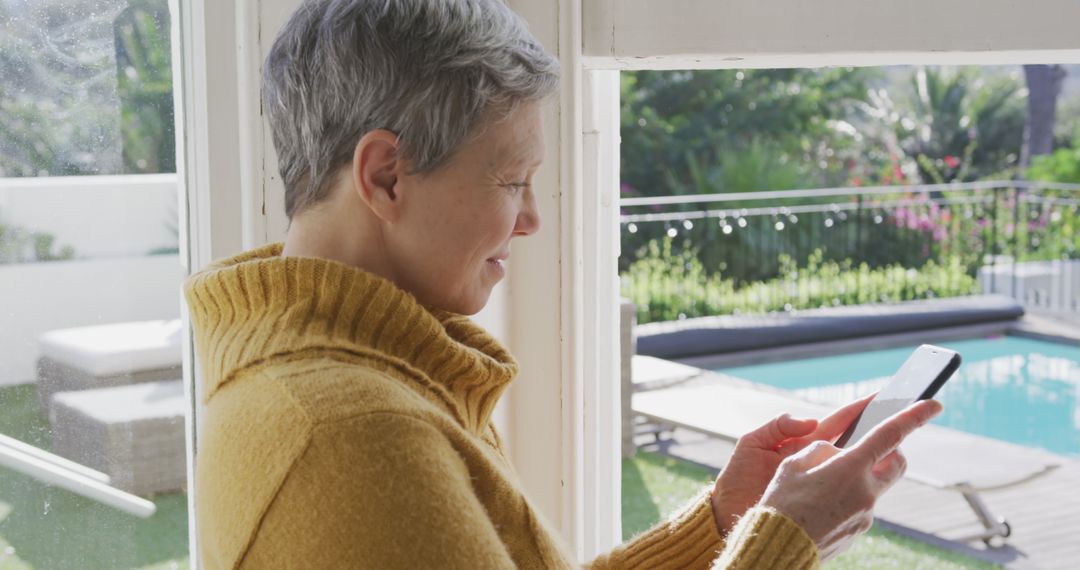 Senior Woman Using Smartphone by Window Overlooking Garden Pool - Free Images, Stock Photos and Pictures on Pikwizard.com