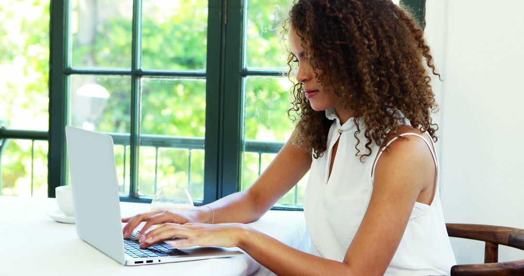 Young African American woman using a laptop at a window-side desk in daylight - Free Images, Stock Photos and Pictures on Pikwizard.com