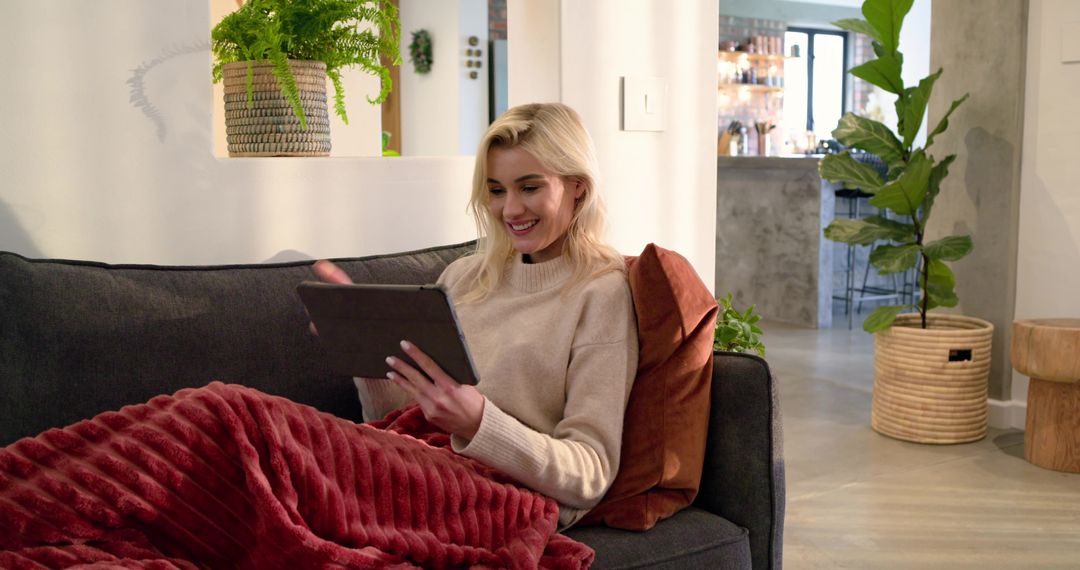 Young woman relaxing on sofa using tablet under plush red blanket in modern living room