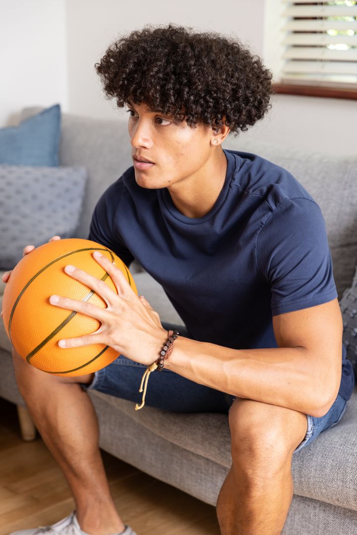 Athletic young man holding basketball on sofa preparing for pickup game with focused expression