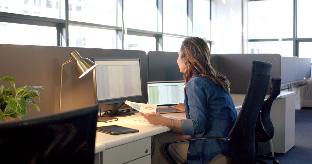Professional woman analyzing data on dual monitors in bright modern open-plan office