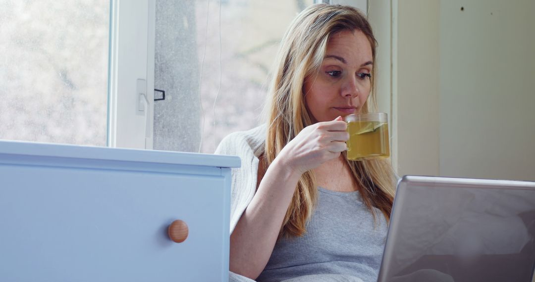 Woman Enjoying Herbal Tea While Using Laptop Near Window - Free Images, Stock Photos and Pictures on Pikwizard.com