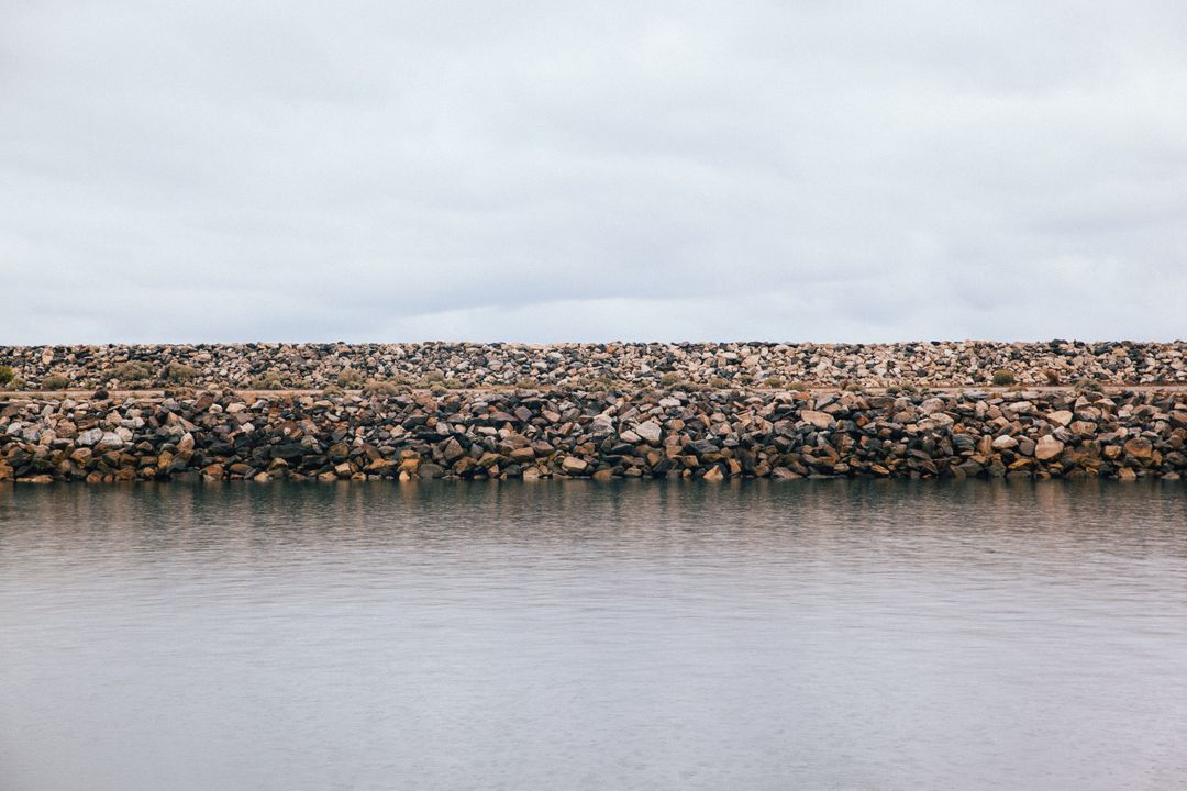 Calm Water Surface with Stone Shoreline on Cloudy Day - Free Images, Stock Photos and Pictures on Pikwizard.com