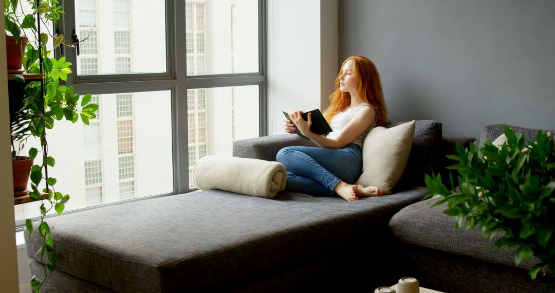 Redhead Woman Relaxing on Sofa Reading Book by Large Window - Free Images, Stock Photos and Pictures on Pikwizard.com