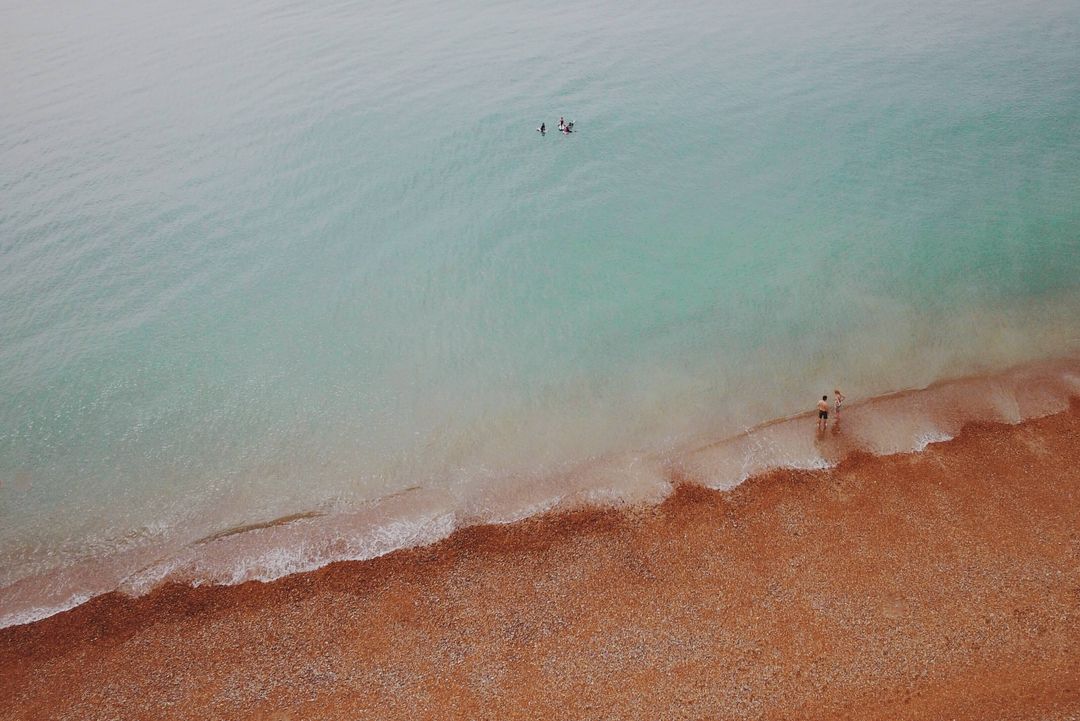 Aerial turquoise sea meeting pebbled red beach with people wading and paddling