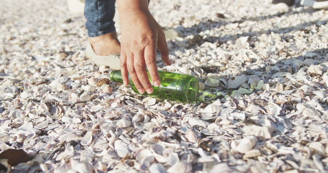 Hand Picking Up Green Bottle from Seashell-Covered Beach Shore - Free Images, Stock Photos and Pictures on Pikwizard.com