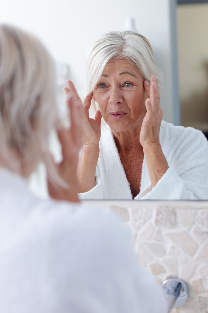 Vertical image of smiling mature caucasian woman inspecting her face in bathroom mirror, copy space - Free Images, Stock Photos and Pictures on Pikwizard.com