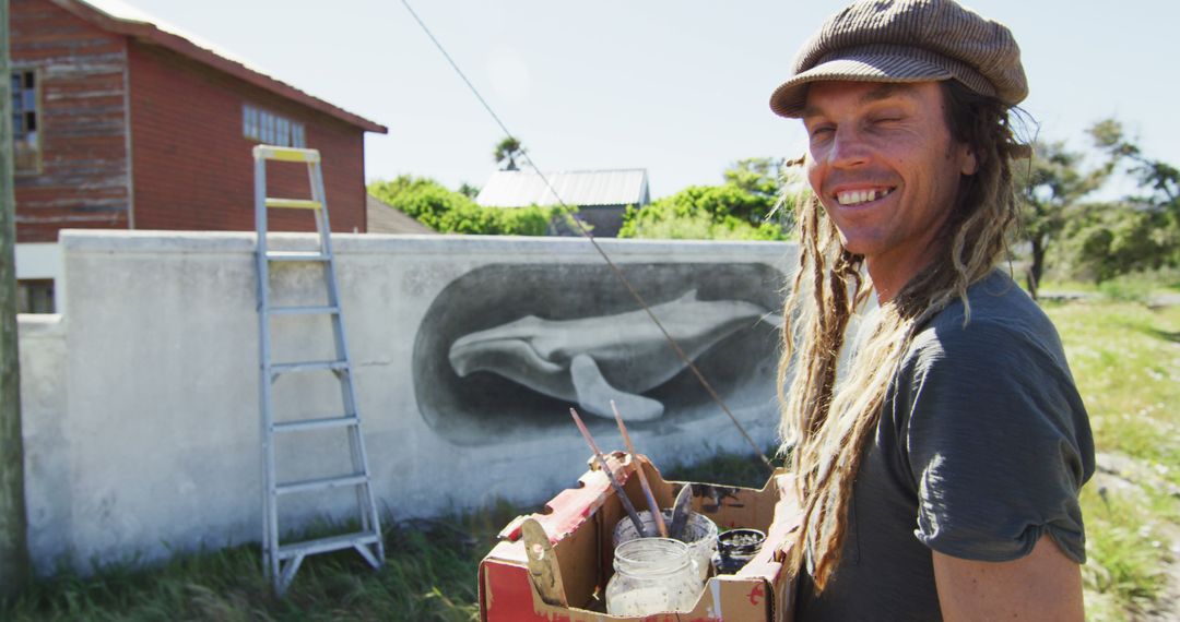 Image of caucasian male artist with dreadlocks holding paints and smiling in front of mural on wall - Free Images, Stock Photos and Pictures on Pikwizard.com