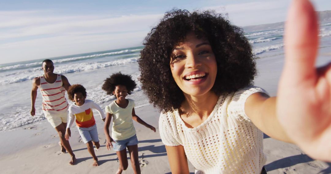 Smiling woman taking selfie with family running and laughing on sunny beach shoreline