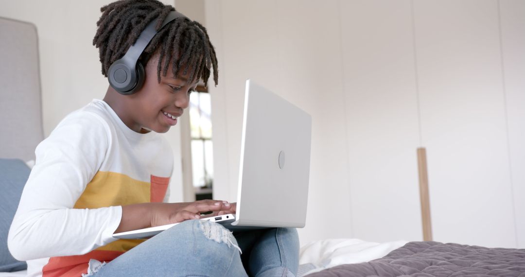 Smiling Boy With Headphones Using Laptop on Bed at Home
