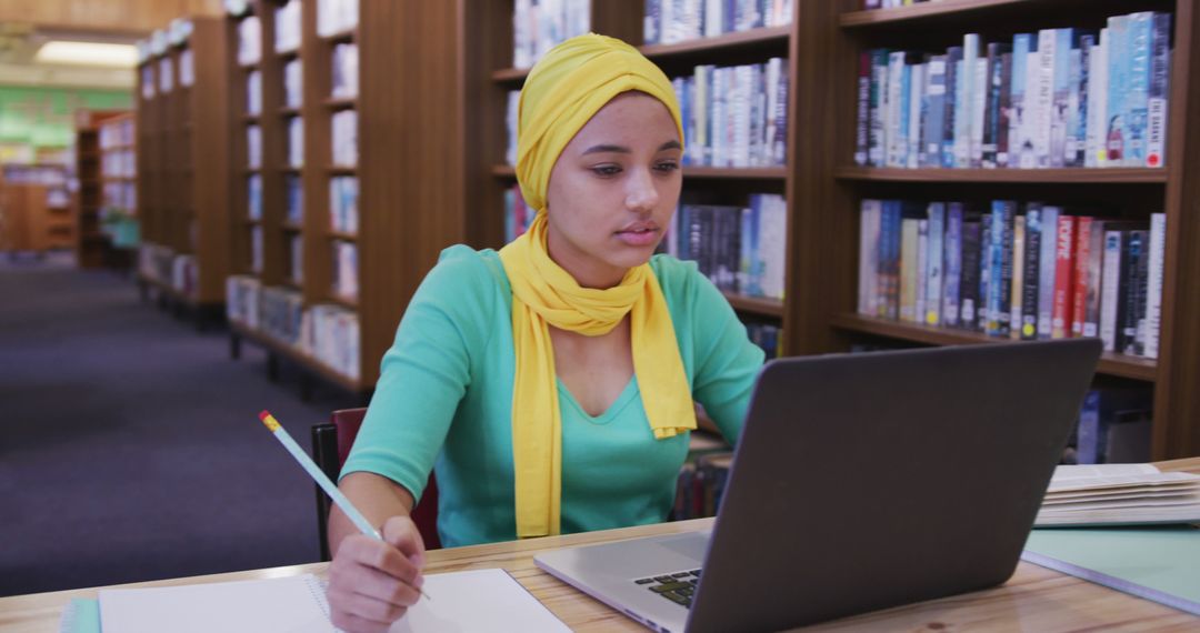 Focused Young Woman with Yellow Headscarf Using Laptop in Library - Free Images, Stock Photos and Pictures on Pikwizard.com