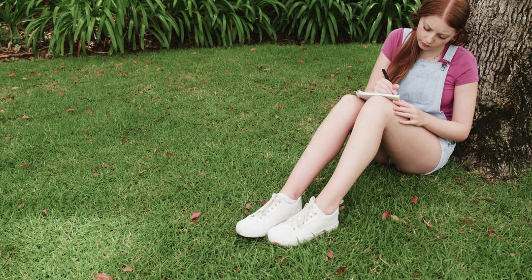 Young woman journaling under tree on green lawn with ample copy space for text