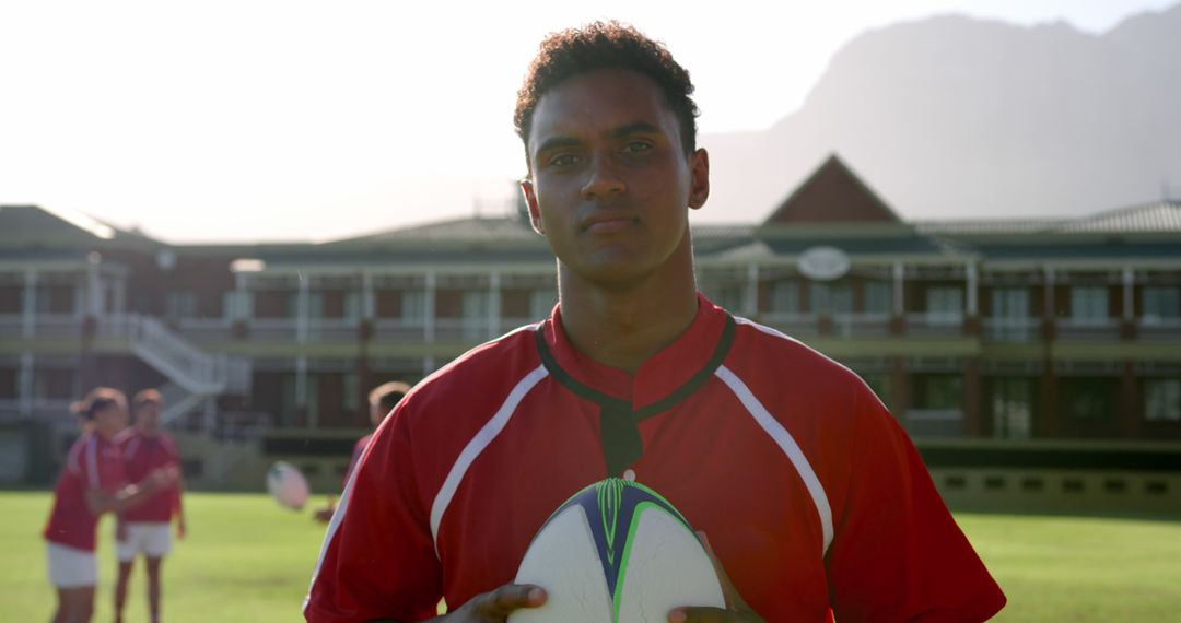 Confident Rugby Player Holding Ball on Field at Sunrise with Teammates in Background - Free Images, Stock Photos and Pictures on Pikwizard.com