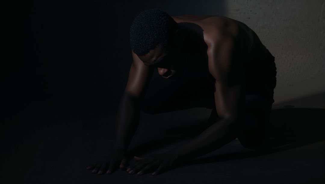 Low-key moody portrait of kneeling shirtless man with sculpted shoulders and deep shadows