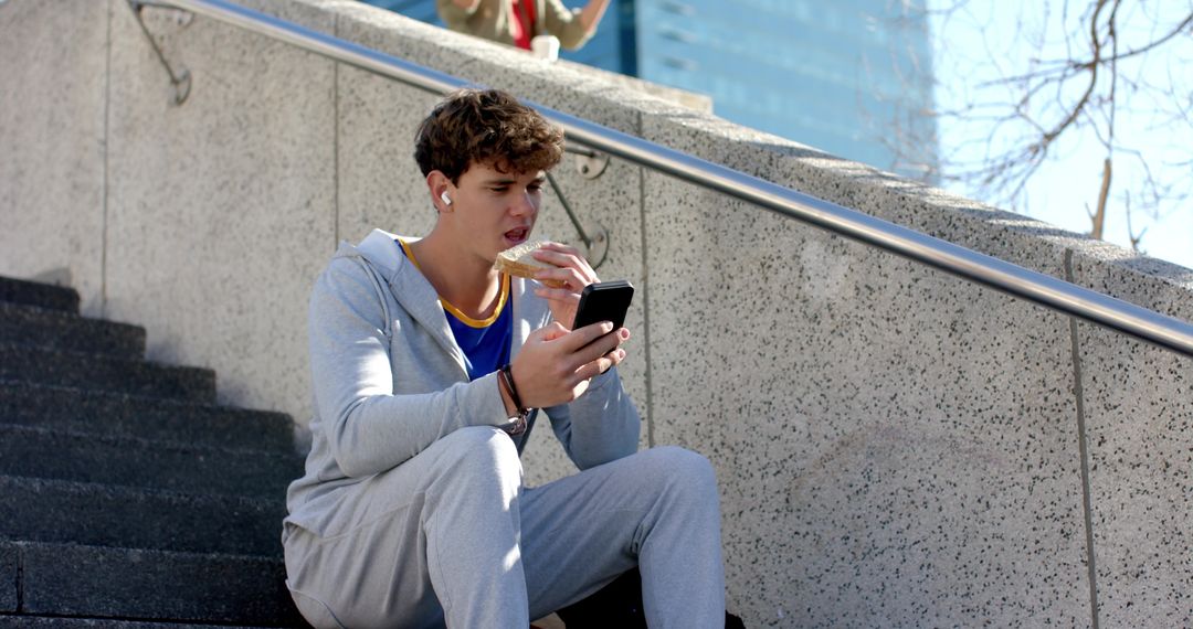 Young man eating sandwich while checking smartphone on urban steps with earbuds