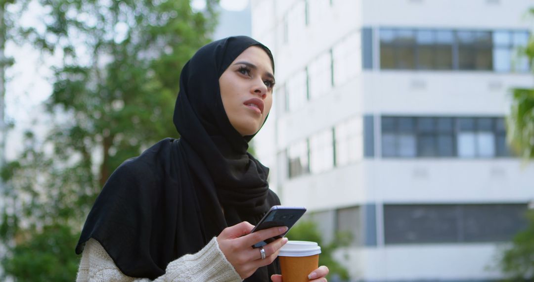 Young Muslim Woman Holding Coffee and Smartphone Outdoors - Free Images, Stock Photos and Pictures on Pikwizard.com