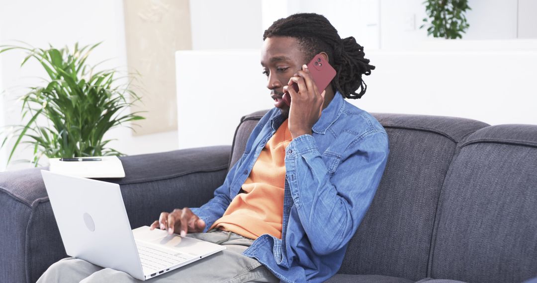 African American man working from home on sofa using laptop and smartphone