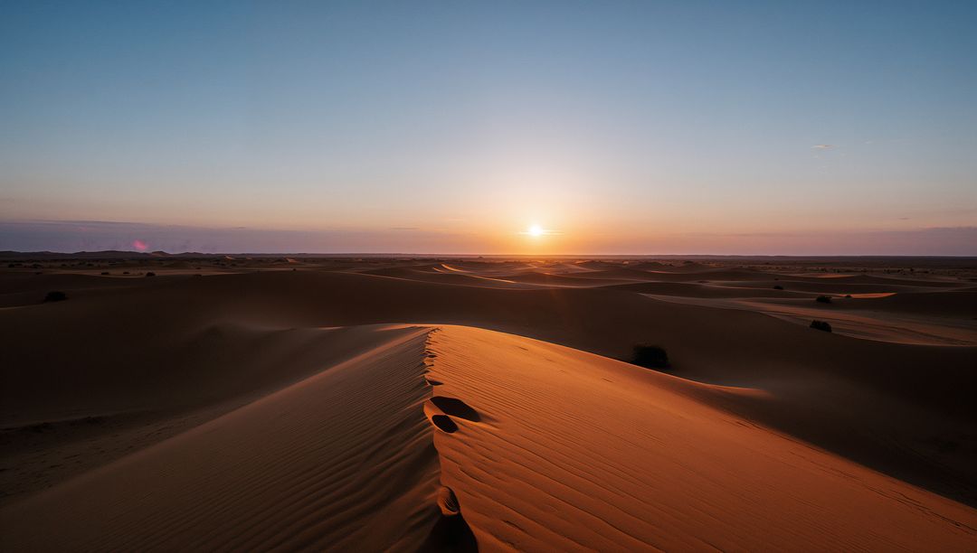 Sunlit dune ridge glowing at sunset revealing ripple patterns and lone footprints over vast desert