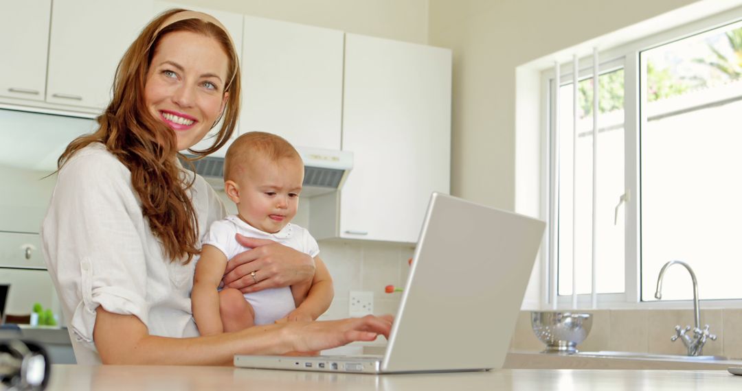 Smiling Mother Working Laptop Holding Baby in Kitchen - Free Images, Stock Photos and Pictures on Pikwizard.com