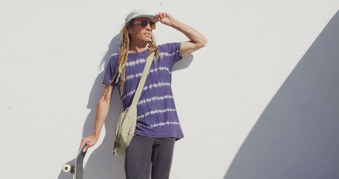 Image of caucasian man with dreadlocks holding skateboard leaning against wall in the sun - Free Images, Stock Photos and Pictures on Pikwizard.com