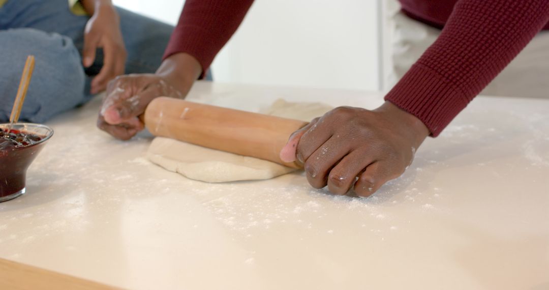 Hands of diverse couple rolling dough on kitchen counter with wooden rolling pin and jam