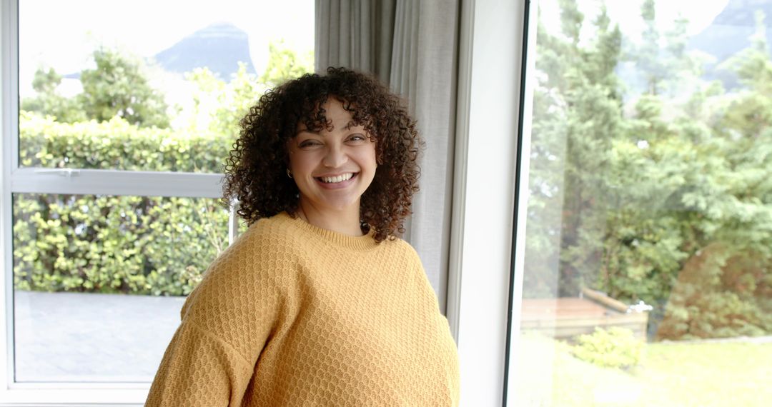 Smiling woman in mustard sweater by large windows enjoying natural light and garden view