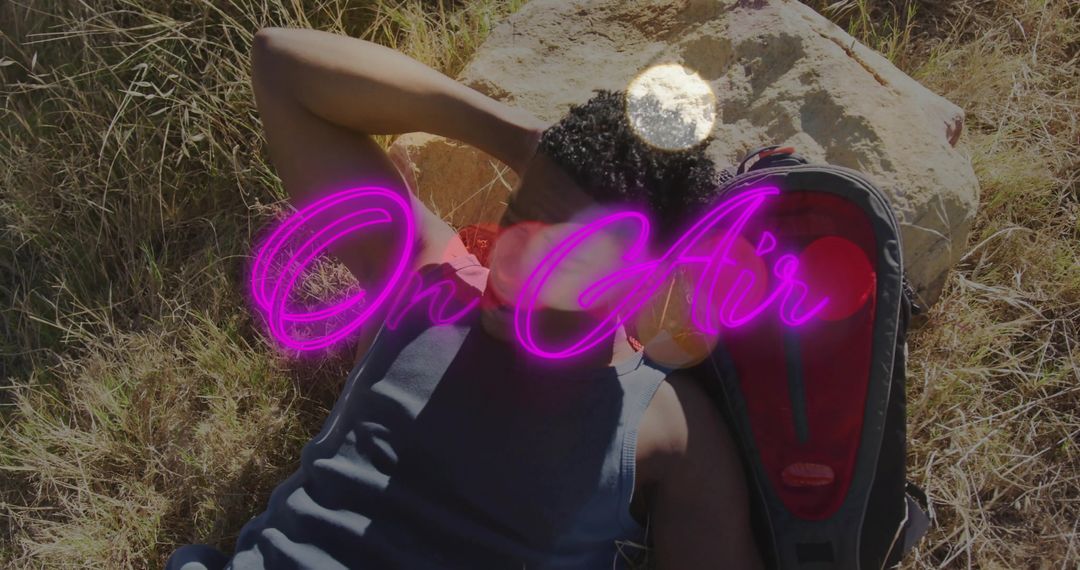 Reclining man wearing tank top resting on grass beside boulder with backpack and neon On Air overlay