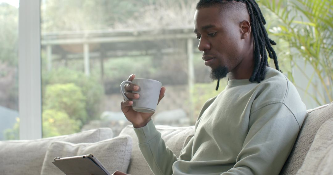 African American man relaxing on sofa holding mug and scrolling tablet by bright window