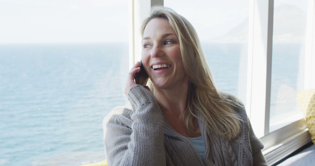 Woman Chatting on Phone by Seaside Window, Smiling Relaxedly - Free Images, Stock Photos and Pictures on Pikwizard.com