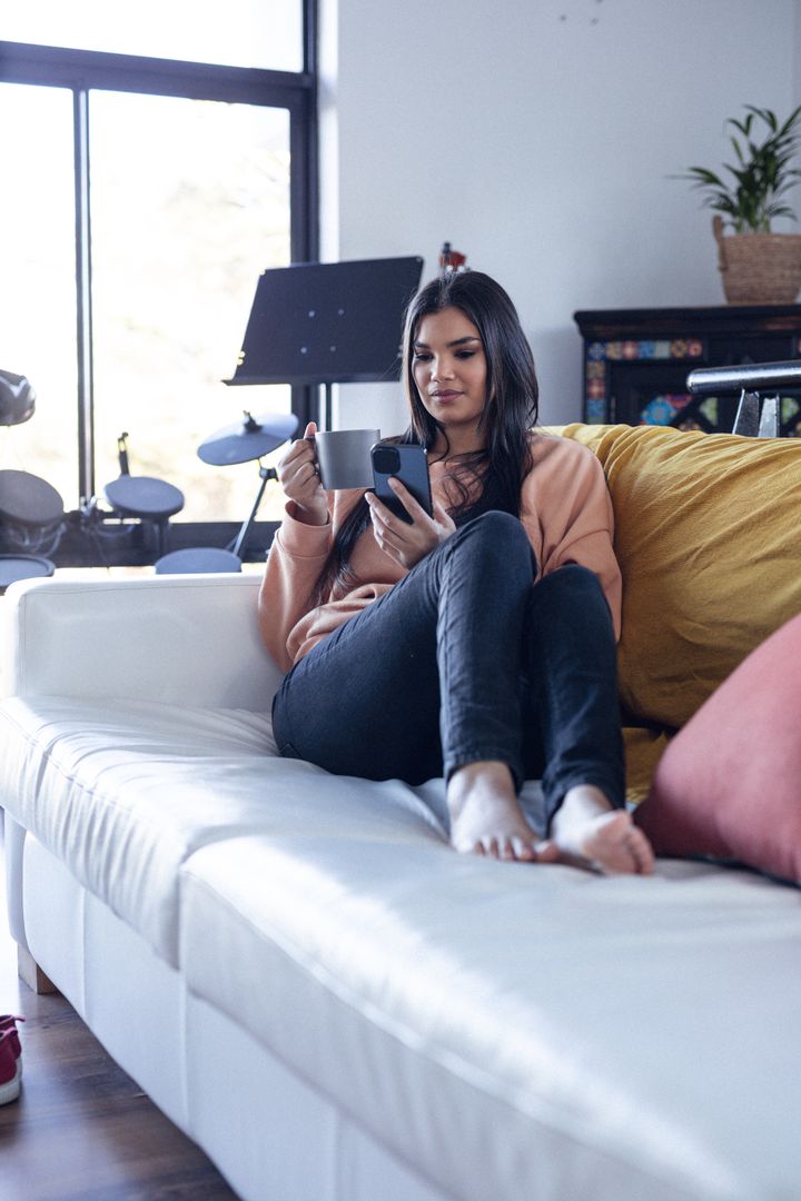 Vertical image of relaxed biracial woman sitting on sofa with coffee and smartphone - Free Images, Stock Photos and Pictures on Pikwizard.com