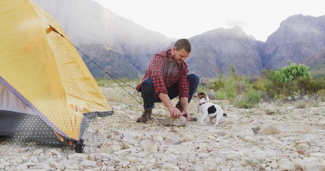 Man Securing Tent Near Mountainside with Dog