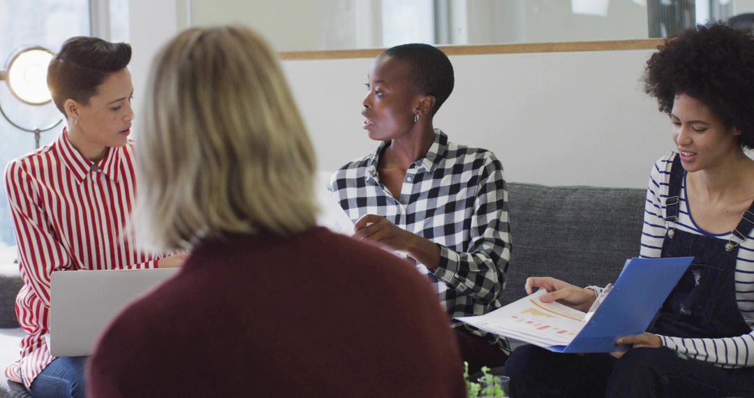Diverse Female Team Collaborating in Modern Office Lounge Sharing Laptop, Charts and Binder