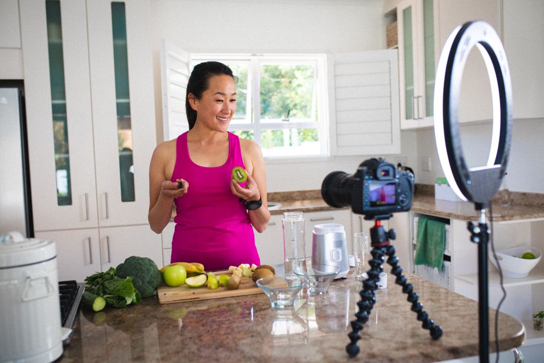 Asian female vlogger with fruits smiling at camera while making healthy juice at home - Free Images, Stock Photos and Pictures on Pikwizard.com
