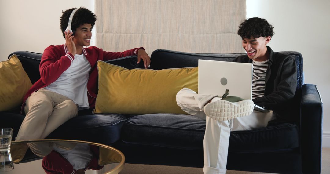 Young men relaxing on sofa sharing laptop and headphones at home for casual collaboration