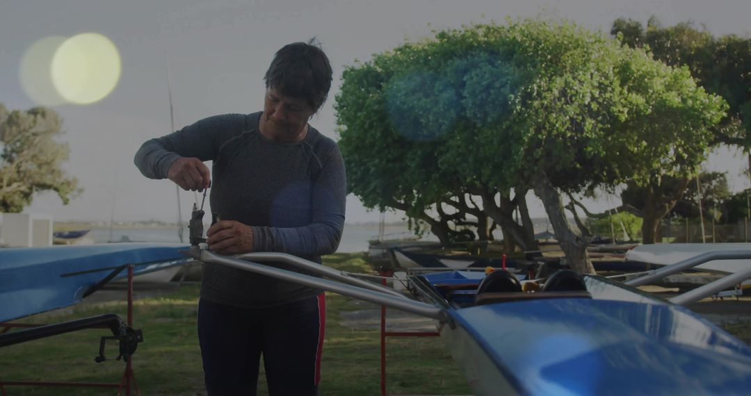 Adjusting rigger on blue rowing shell by experienced woman at waterfront boatyard
