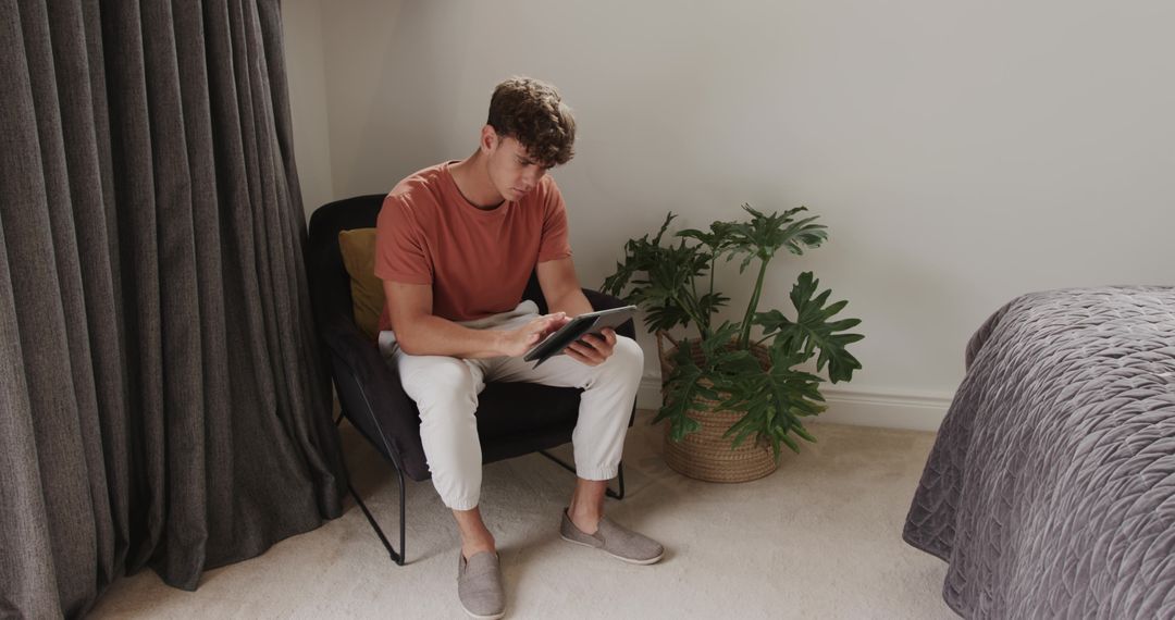 Young man using tablet with stylus in minimalist bedroom chair surrounded by plant