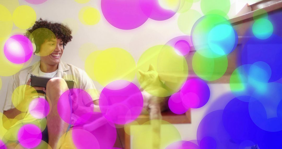 Young man relaxing on stairs holding tablet with cat companion amid colorful bokeh overlay