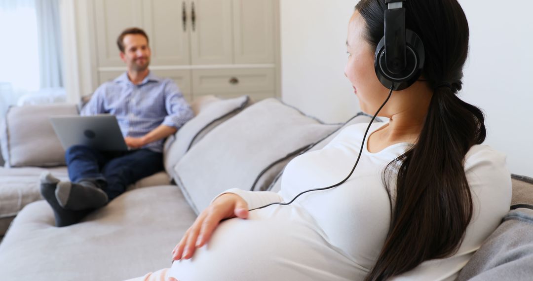 Expectant Mother Relaxing with Headphones on Couch, Partner Working in Background - Free Images, Stock Photos and Pictures on Pikwizard.com