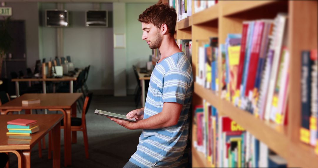 Young Man Using Tablet in Library Leaning Against Bookshelf - Free Images, Stock Photos and Pictures on Pikwizard.com